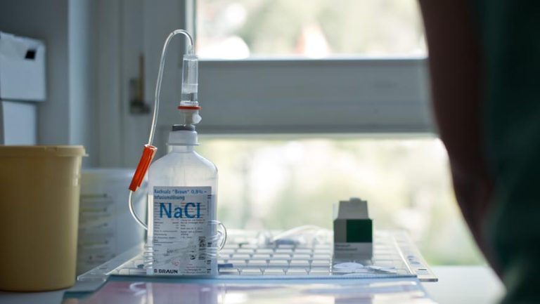 Bild zeigt eine Flasche Natrium-Chlorid-Lösung mit Versorgungsschlauch und einer Medikamentendose