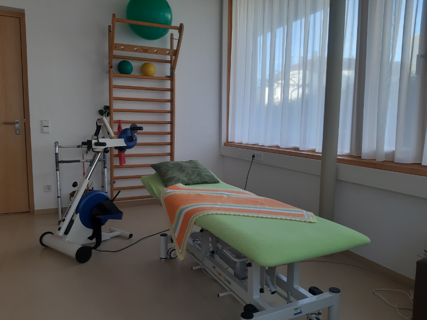 Auf dem Bild ist der Therapieraum der Tiroler Hospiz Gemeinschaft zu sehen. Es ist eine Liege, mehrere Trainingsgeräte und eine Sprossenwand mit Bällen zu sehen.