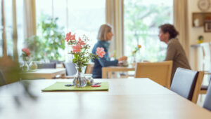 Im Bild ist ein Tisch im Hospizcafé mit Blumenschmuck im Hintergrund sind zwei Personen zu sehen.