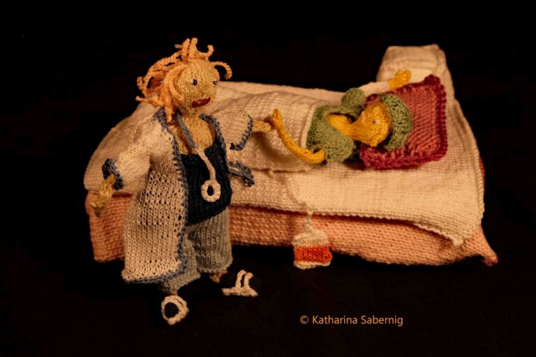 Zwei gestrickte Puppen in einer Szene aus dem Krankenhaus. Links steht eine Ärztin oder Pflegekraft mit wirrem, blondem Haar, weißem Kittel und Stethoskop. Rechts liegt eine Patientin im Bett, zugedeckt mit einer rosa-weißen Decke, ein grünes Mützchen auf dem Kopf. Eine Infusion hängt vom Bett herunter. Die Szene ist komplett aus Wolle gefertigt und mit viel Liebe zum Detail gestaltet. Hintergrund ist dunkel, Fokus liegt auf den Figuren.