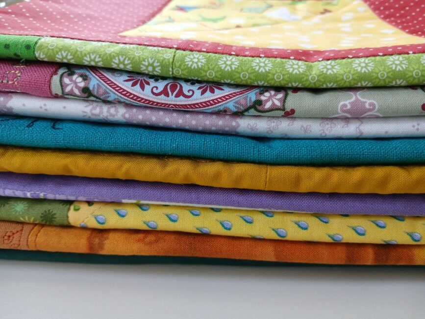 Ein Stapel bunter, sorgfältig gefalteter Stoffstücke oder Quilts liegt aufeinander. Die Stoffe zeigen verschiedene Farben und Muster, darunter Punkte, Blumen, Tropfen und Ornamente in Gelb, Orange, Lila, Grün, Türkis und Rot. Die Nahaufnahme betont die Vielfalt und Struktur der Textilien.