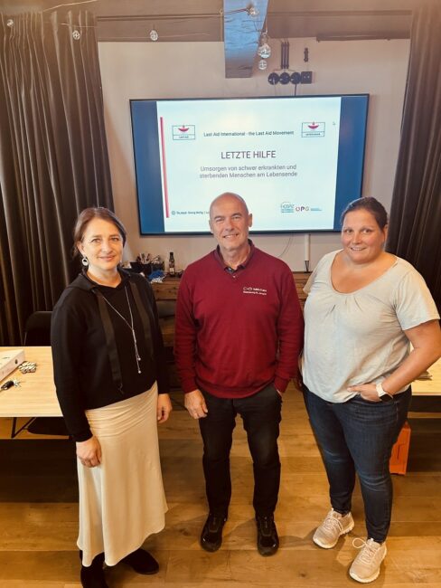 3 Personen stehend, im Hintergrund ein Bildschirm an der Wand