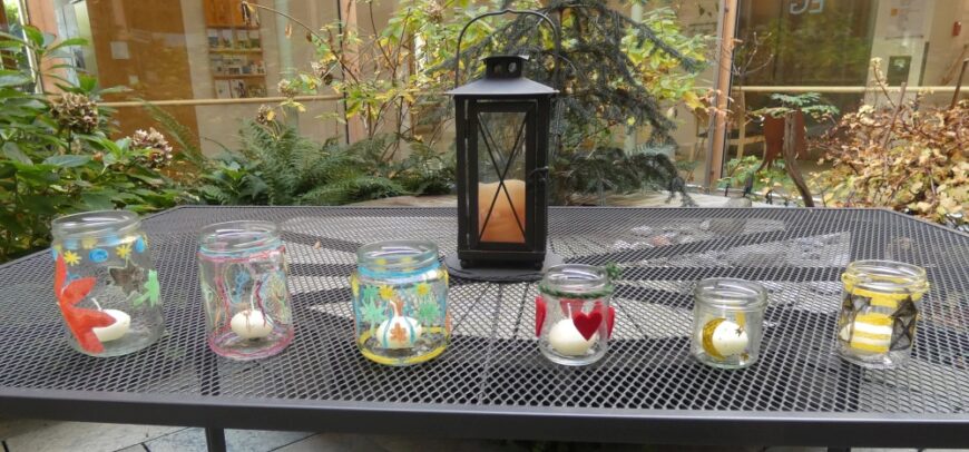 Sechs gebastelte Martinslaternen aus Marmeladengläsern stehen auf einem schwarzen Metalltisch im Freien. Jedes Glas ist individuell mit Farben und Motiven wie Herzen, Sternen, Monden und abstrakten Mustern verziert und enthält ein kleines Teelicht. In der Mitte steht eine hohe, schwarze, schmiedeeiserne Laterne mit einer brennenden Kerze. Im Hintergrund ist ein Innenhof mit herbstlicher Bepflanzung zu erkennen.