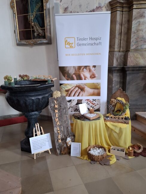 Eine Gedenk- und Informationsstation der 'Tiroler Hospiz Gemeinschaft' in einer Kapelle. Im Hintergrund steht ein großes Banner mit dem Logo und dem Slogan 'WIR BEGLEITEN MENSCHEN'. Davor sind mehrere Tische mit gelben Tüchern arrangiert. Links steht ein großes, schwarzes Steingefäß, das mit Kerzen und getrockneten Blumen geschmückt ist. In der Mitte steht ein langes Stück Baumrinde mit den vertikal eingravierten Buchstaben 'INNEHALTEN'. Rechts liegt in einem Korb eine große Anzahl weißer, runder Steine. Ein Schild auf der linken Seite erklärt die Bedeutung der Steine und lädt zur Interaktion ein. Rechts außen steht ein hölzerner Thron mit der Aufschrift 'TRÖSTEN' auf einem gelben Tuch.