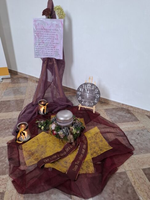 Eine Gedenkecke mit einer Urne und Symbolen der Zeit. Im Zentrum liegt eine silberne Urne, umgeben von einem Kranz aus getrockneten Blumen. Die Urne steht auf dunklem und gelbem Stoff und zwei Schleifenbändern mit der Aufschrift 'Der Liebe gehören Zeit und Tränen' und 'Dem Tod gehört nur ein Augenblick'. Dahinter steht eine Staffelei mit einem beschriebenen Leinwandbild. Rechts daneben steht eine kleine, runde Uhr auf einer Staffelei, deren Zeiger auf kurz vor zwölf stehen. Zwei Teelichter in Gläsern flankieren die Anordnung
