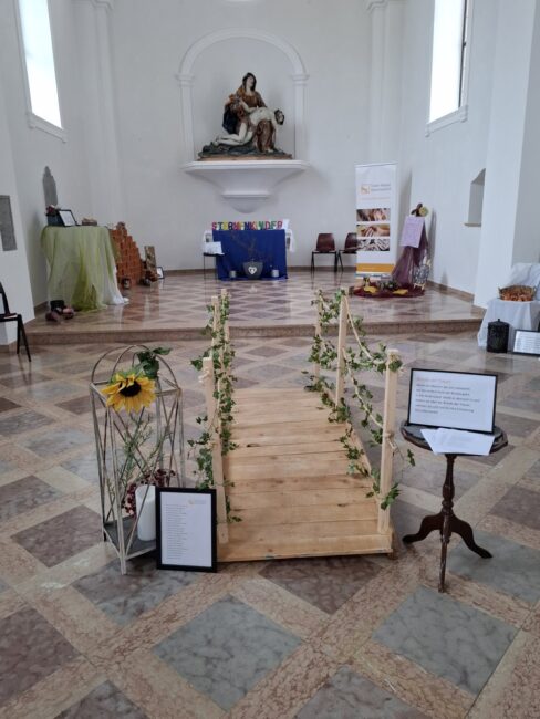 Gesamtansicht eines hellen Trauerraums, der mit verschiedenen Gedenkinstallationen gestaltet ist. Im Vordergrund steht eine kleine, begehbare Holzbrücke, deren Geländer mit Efeu und Seilen verziert ist. Links neben der Brücke befindet sich eine weiße Laterne mit einer Sonnenblume und einer Kerze. Rechts steht ein kleiner Tisch mit einem Schild, das den Text 'Brücke zur Erinnerung' trägt. Im erhöhten Altarbereich im Hintergrund sind verschiedene Stationen zu sehen, darunter die Installation 'Sternenkinder', eine Mauer aus Ziegelsteinen (Klagemauer) und in der Mitte eine Figurengruppe auf einem Sockel."