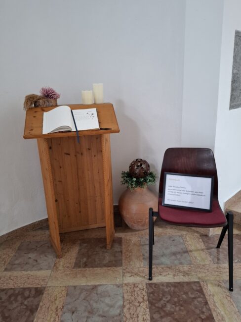 Ein schlichtes, hellbraunes Holzpult trägt ein aufgeschlagenes Gästebuch und drei weiße Stumpenkerzen. Rechts daneben steht ein brauner Stuhl, auf dessen Sitzfläche ein gerahmtes Schild liegt. Der Text des Schildes beginnt mit 'Gästebuch' und fordert die Besucher freundlich auf, ihre Gedanken niederzuschreiben. Neben dem Pult steht eine große, bauchige Tonvase, die mit einem Kranz aus getrockneten Materialien und einer braunen Metallkugel dekoriert ist.