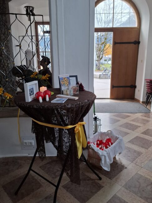 Ein runder Stehtisch im Eingangsbereich eines Trauerraums, der mit einem braunen Tuch und einer gelben Schleife dekoriert ist. Auf dem Tisch stehen zwei kleine, rote Keramikpilze, ein brennendes Teelicht und gerahmte Informationen. Rechts neben dem Tisch steht ein weißer Korb, gefüllt mit kleinen roten Objekten (vermutlich Glückspilze oder Herzen) zum Mitnehmen ('Give-aways'). Im Hintergrund ist eine große, offene Holztür mit Blick nach draußen auf einen Baum mit gelben Blättern zu sehen.