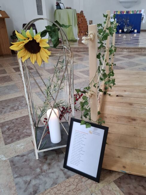 Eine Gedenkecke mit einer großen, weißen Rostlaterne und einer hölzernen Brücke. Die Laterne enthält eine dicke weiße Kerze und ist mit einer großen gelben Sonnenblume, Beeren und grünen Zweigen dekoriert.