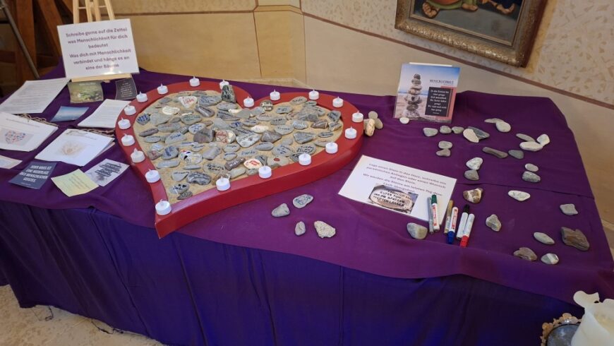 Ein großer Tisch, der mit einem violetten Tuch bedeckt ist, dient als Station zum Thema Menschlichkeit. Im Zentrum liegt ein großes, rotes Holzherz, das mit Sand gefüllt und mit vielen kleinen Steinen bedeckt ist, auf denen Botschaften geschrieben stehen. Um das Herz herum stehen mehrere kleine weiße Kerzen. Links und rechts liegen Informationsblätter und weitere unbeschriftete Steine sowie Filzstifte, mit denen Besucher ihre eigenen Wünsche oder Gedanken auf die Steine schreiben können. Ein Schild fordert die Besucher auf: 'Schreibe gerne auf die Zettel, was Menschlichkeit für dich bedeutet, was dich mit Menschlichkeit verbindet und hänge es an eine der Bäume'.