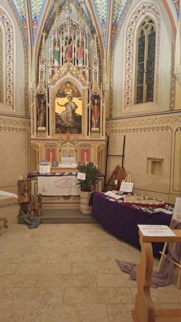 Der Innenraum einer Kapelle, fokussiert auf den Altarraum. Im Zentrum steht ein reich verzierter, goldener gotischer Flügelaltar mit einer Darstellung von Christus in der Mitte. Der Altar ist mit einem weißen Tuch und einem roten Textbanner dekoriert, davor stehen Kerzen und eine Pflanze in einem Holzfass. Rechts im Vordergrund steht ein Tisch, der mit einem violetten Tuch abgedeckt ist und verschiedene Gedenkobjekte und Schilder trägt, darunter eines mit der Aufschrift 'Trauergruppe Sebastianikapelle'. Die Wände und Bögen sind mit kunstvollen, farbigen Mustern und Malereien verziert.