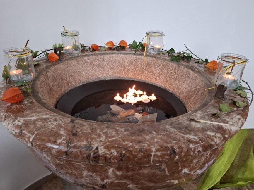 Ein großer, runder Brunnen oder Taufstein aus rotbraun marmoriertem Naturstein. Auf der dunklen Wasseroberfläche schwimmen mehrere kleine Kerzen, die hell leuchten. Der Rand des Steingefäßes ist mit Efeuranken, Physalis (Lampionblumen) und vier weiteren Kerzen in kleinen Gläsern dekoriert."
