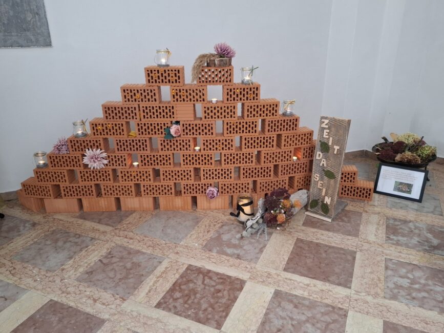 Eine pyramidenförmig aufgeschichtete Mauer aus roten Lochziegeln, die mit Kerzen und Blumen geschmückt ist. In den Löchern der Ziegel stecken kleine, zusammengerollte Zettel. Rechts daneben lehnt ein Holzbrett mit der vertikalen Aufschrift 'ZEIT DA SEIN'.