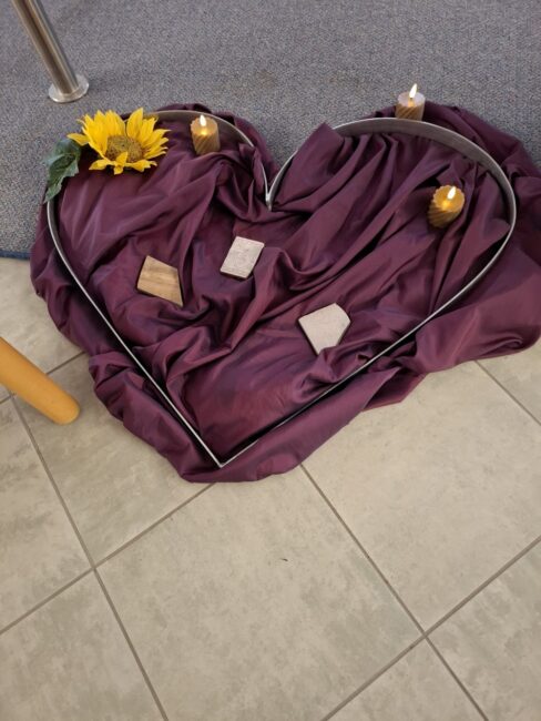 Ein symbolisches Herz, gefertigt aus einem silbernen Metallrahmen, liegt auf einem hell gefliesten Boden. Das Innere des Herzens ist mit einem dunkelvioletten, weich fallenden Stoff ausgelegt. Darauf liegen einige helle, unregelmäßige Steine und Holzstücke. Das Arrangement ist mit drei wachsfarbenen Stumpenkerzen und einer gelben Sonnenblume dekoriert, was eine Gedenkstätte darstellt.