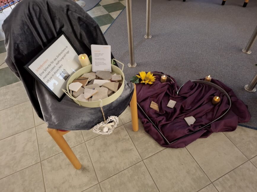 Ein großes, auf dem Boden liegendes Herz, das von einem silbernen Metallrahmen umgeben ist. Das Innere des Herzens ist mit einem dunkelvioletten, weich fallenden Stoff ausgelegt. Auf dem Stoff liegen mehrere kleine, helle Holzstücke und Steine. Das Arrangement ist mit drei wachsfarbenen Kerzen in Stumpenform und einer künstlichen Sonnenblume dekoriert. Der Boden besteht aus hellen, quadratischen Fliesen.