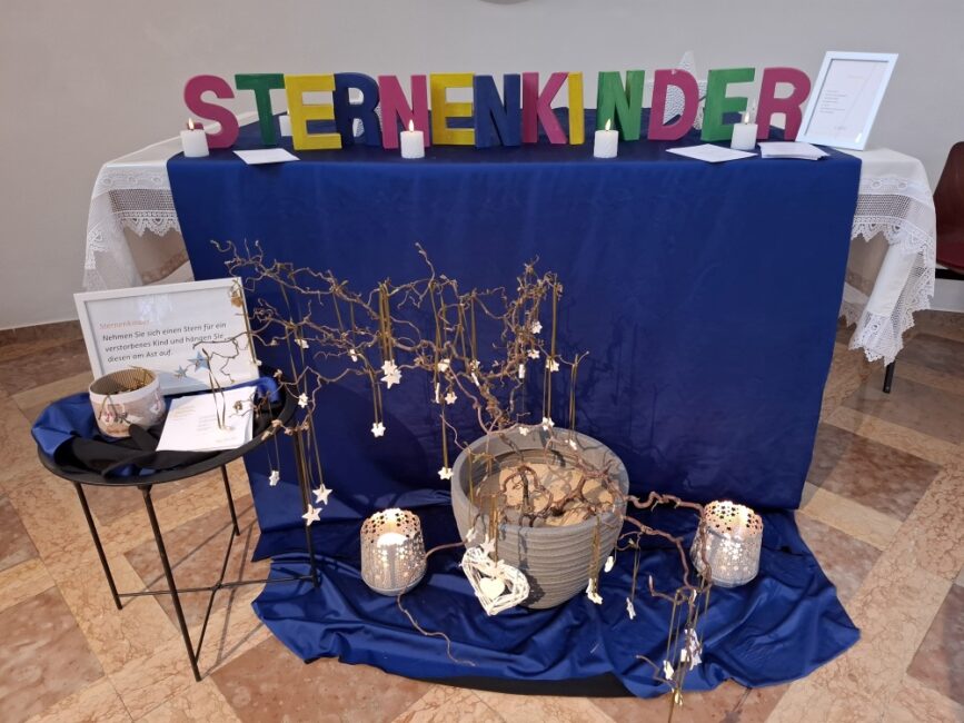 Ein Gedenktisch, der den 'Sternenkindern' gewidmet ist. Auf einem blau abgedeckten Tisch stehen große, bunte Holzbuchstaben, die das Wort 'STERNENKINDER' bilden. Davor steht eine große graue Schale mit Ästen, von denen kleine weiße Sterne und Herzen herabhängen. Auf dem Boden um die Schale liegen blaue Tücher und zwei Kerzen in silbernen Windlichtern. Links steht ein Beistelltisch mit einem Schild, das die Besucher auffordert, einen Stern für ein verstorbenes Kind aufzuhängen.