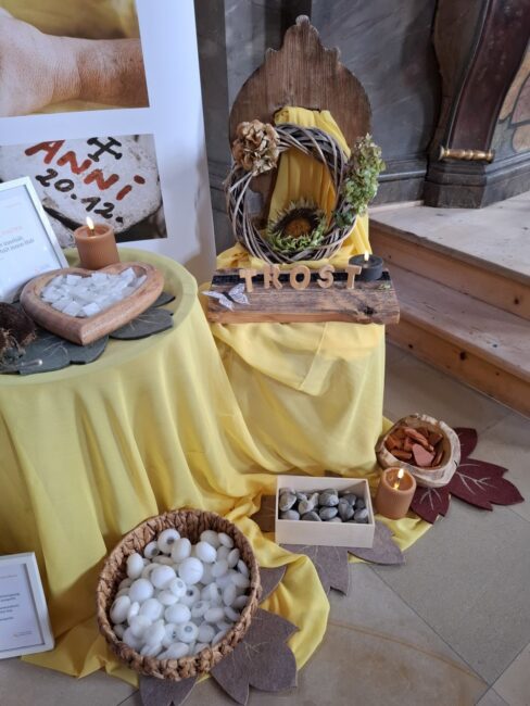 Detailansicht einer Gedenkstation in warmen Gelbtönen, thematisch auf Trost ausgerichtet. Im Zentrum liegt ein kleines Holzbrett mit dem Schriftzug 'TROST'. Darüber hängt ein Kranz aus Weidenzweigen, verziert mit getrockneten Blumen und einer Sonnenblume. Der Bereich steht auf einem Tisch mit einem leuchtend gelben Tuch. Links auf dem Tisch liegt ein herzförmiges Gefäß mit kleinen weißen, würfelförmigen Objekten (Steine). Auf dem Boden stehen ein Korb mit runden, weißen Steinen und eine kleine Holzkiste mit grauen Kieseln, umgeben von braunen Herbstblättern aus Filz und Kerzen.