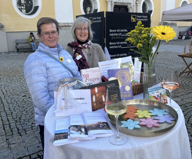 Zwei lächelnde Frauen stehen an einem Informationsstand der Tiroler Hospiz-Gemeinschaft auf einem gepflasterten Platz vor einer Kirche. Auf dem runden, weiß gedeckten Tisch liegen Broschüren, ein Buch mit dem Titel „99 Fragen an den Tod“, bunte Papierblumen in einer Metallschale und Gläser mit Wein. Im Hintergrund ist eine Tafel mit der Aufschrift „Bevor ich sterbe, möchte ich...“ zu sehen.