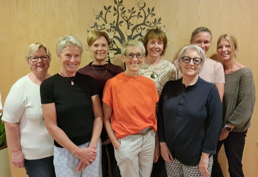 Gruppenfoto von sieben lächelnden Frauen, die nebeneinander vor einer Holzwand mit einem Baum-Wandtattoo stehen.