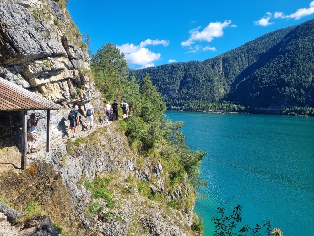 Wandergruppe auf einem schmalen Pfad an einer Felswand oberhalb eines türkisblauen Sees mit bewaldeten Bergen im Hintergrund.