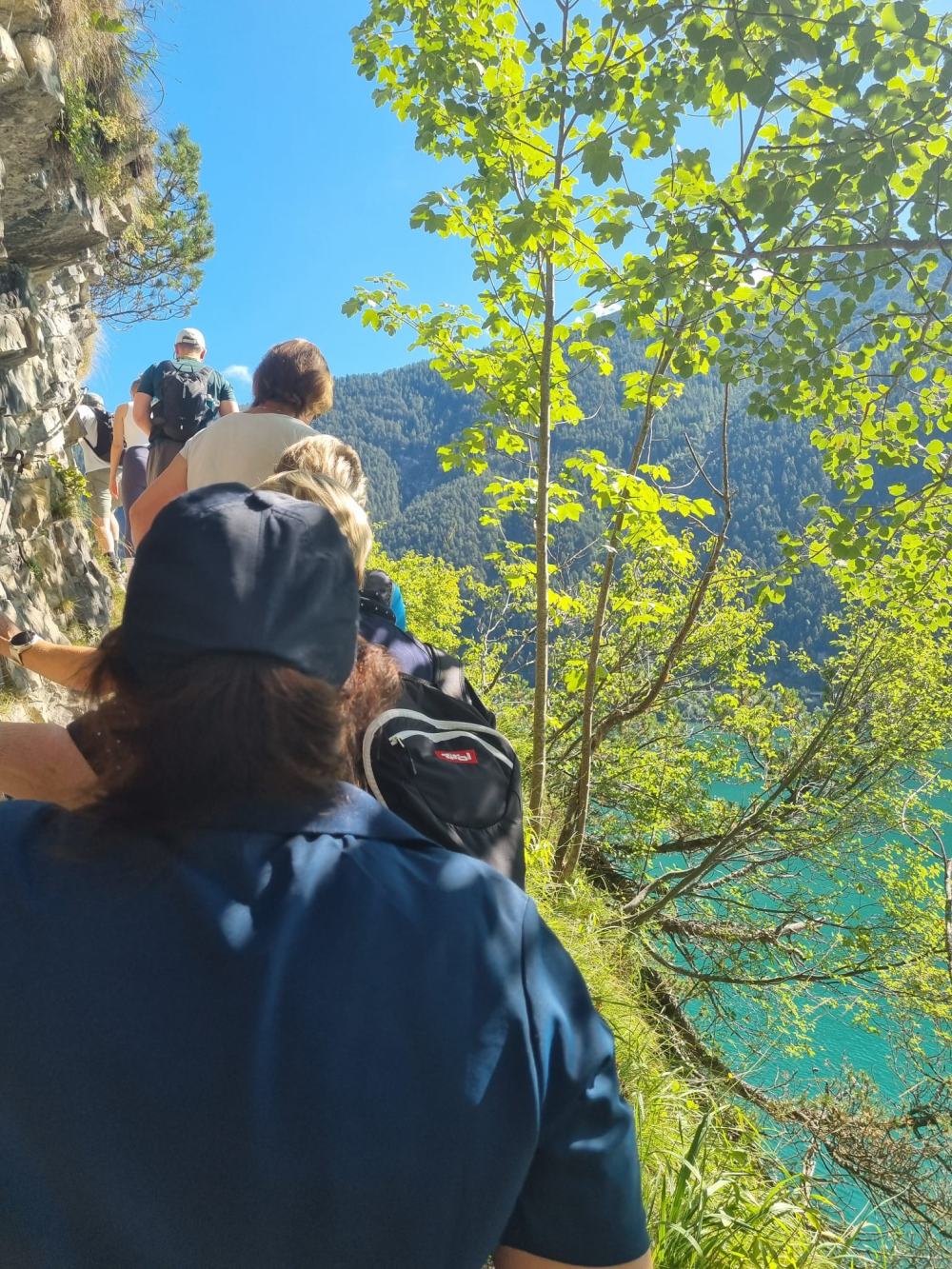 Wandergruppe hintereinander auf einem schmalen Pfad an einer Felswand oberhalb eines türkisblauen Sees.
