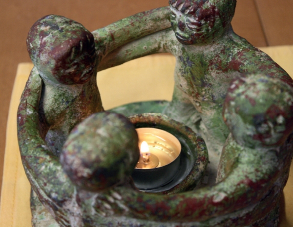 Eine Keramik-Skulptur mit patinaartiger, grün-roter Oberfläche. Vier stilisierte menschliche Figuren stehen im Kreis, halten sich an den Schultern und bilden ein Miteinander. In ihrer Mitte brennt ein Teelicht.