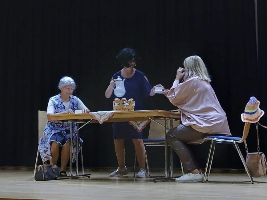 Drei Personen in einem Theaterstück auf einer Bühne. Eine stehende Frau gießt Tee für zwei sitzende Frauen an einem Tisch ein.