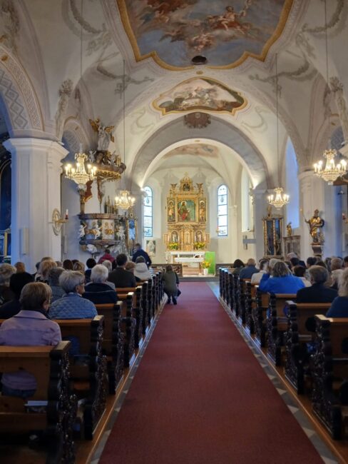 Blick durch den Mittelgang einer voll besetzten, barock verzierten Kirche auf den Altar während eines Gottesdienstes.
