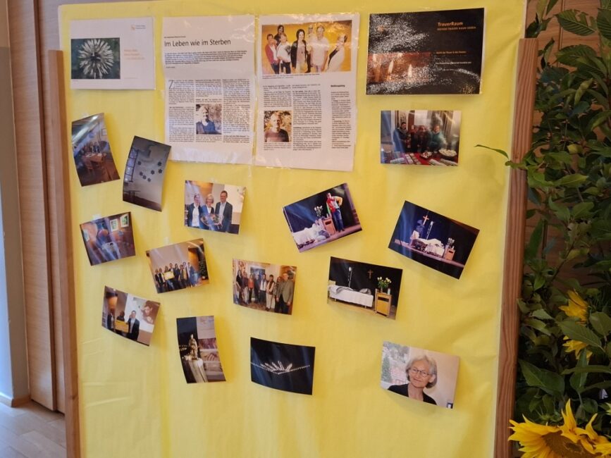 Eine gelbe Infowand in einem Holzrahmen, behängt mit verschiedenen Fotos von Personengruppen, Veranstaltungen, einem Bett und Zeitungsartikeln zum Thema „Hospiz“ und „Trauer“ [cite: 3]. Rechts im Bild stehen eine Grünpflanze und Sonnenblumen[cite: 3].