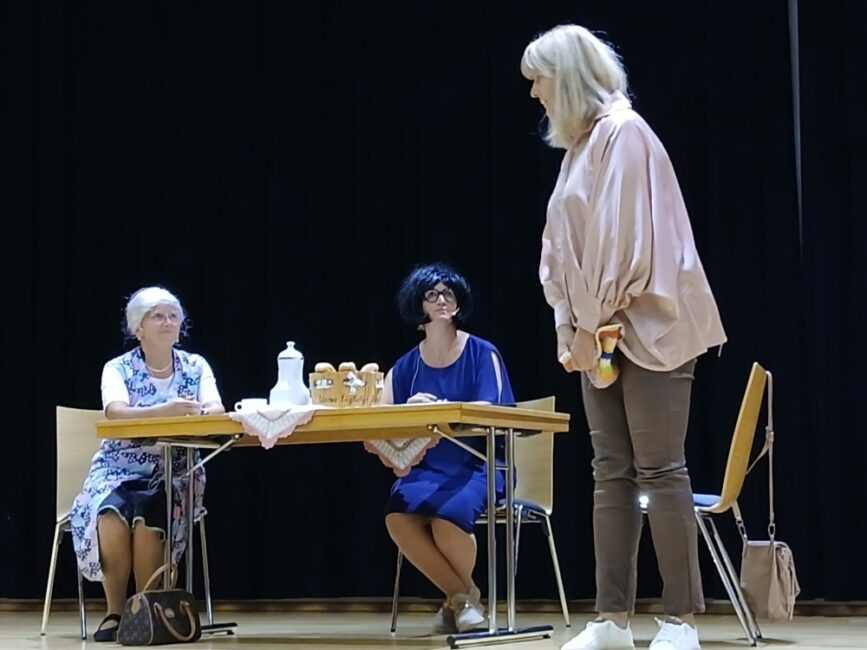 Szene aus einem Theaterstück: Eine stehende Frau mit hellblonder Frisur und rosa Bluse spricht mit zwei Frauen, die an einem Tisch sitzen.