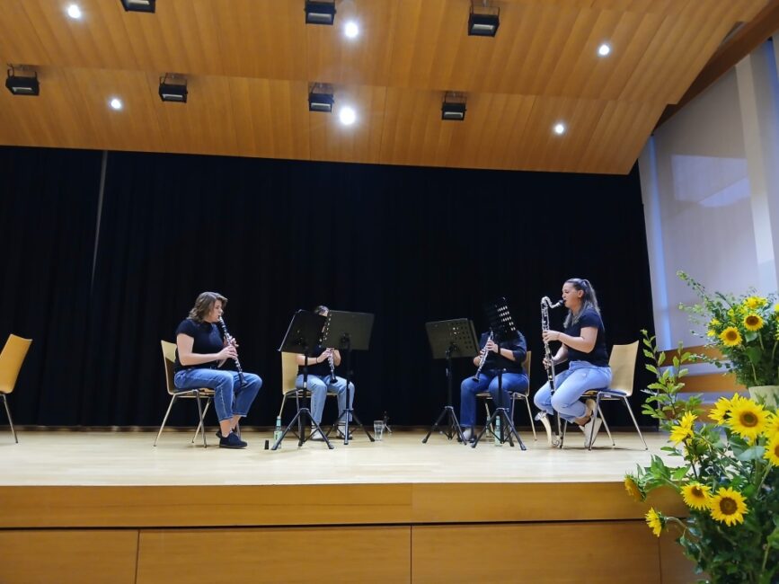 Ein Klarinetten-Quartett aus vier Musikerinnen spielt sitzend auf einer Holzbühne vor Notenständern. Rechts im Vordergrund stehen gelbe Sonnenblumen