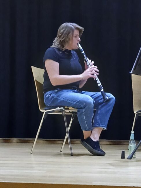 Eine Musikerin mit schulterlangem Haar sitzt auf einem Holzstuhl auf einer Bühne und spielt Klarinette.