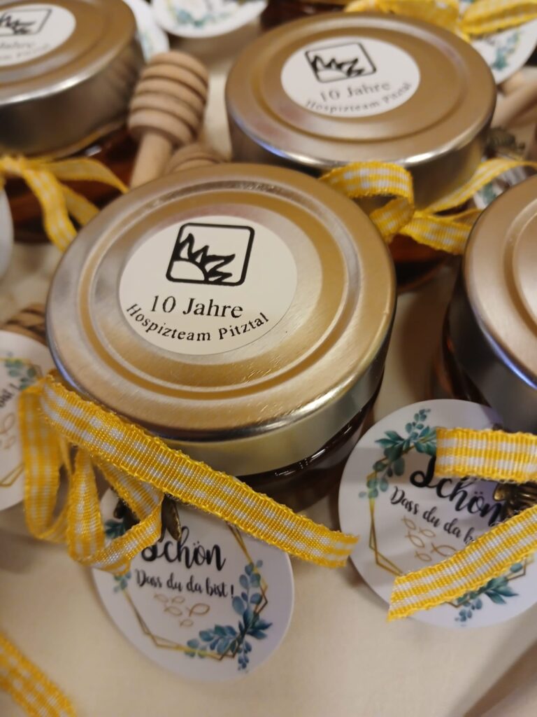 Ein Arrangement aus kleinen Honiggläsern mit goldenen Deckeln. Jedes Glas trägt ein weißes Etikett zum „10 Jahre Hospizteam Pitztal“-Jubiläum und ist mit einer gelb-weiß karierten Schleife und einem Anhänger mit dem Text „schön dass du da bist!“ versehen. Im Hintergrund sind Honigheber aus Holz zu sehen.