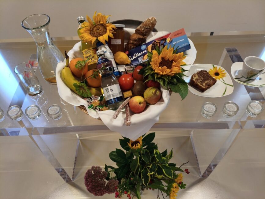 Ein reich gefüllter Geschenkkorb mit Obst, Brot, Süßigkeiten und Sonnenblumen auf einem transparenten Glastisch, flankiert von Glaskaraffen, Kaffeetasse, Kuchen und Teelichtern.