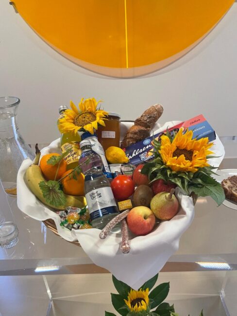 Ein mit einem weißen Tuch ausgelegter Geschenkkorb, gefüllt mit frischem Obst wie Äpfeln, Birnen, Orangen und einer Banane, sowie Tomaten, Getränkeflaschen, Brot, Keksen und Dekoration aus Sonnenblumen.