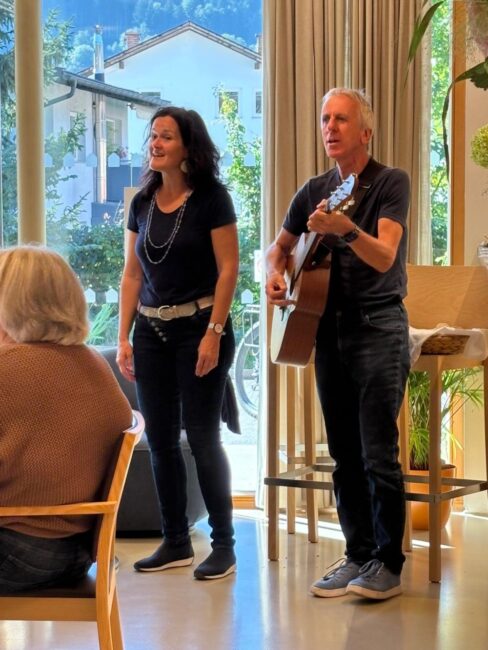 Uschi und Walter Hölbling stehen nebeneinander in einem hellen Raum vor großen Fenstern und singen. Der Mann spielt dazu eine Akustikgitarre, während im Vordergrund im Anschnitt das Publikum zu sehen ist.