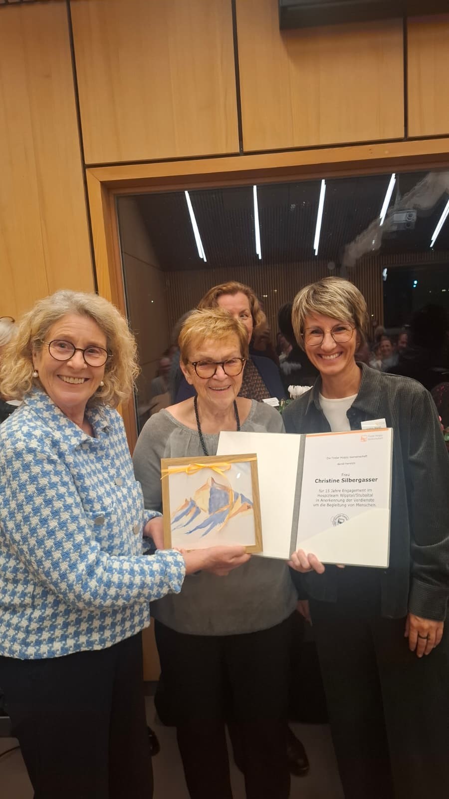 Drei lächelnde Frauen stehen nebeneinander bei einer Ehrung. Die Frau in der Mitte hält ein kleines eingerahmtes Aquarellbild und eine Urkunde der Tiroler Hospiz-Gemeinschaft für Christine Silbergasser für 15 Jahre Engagement.