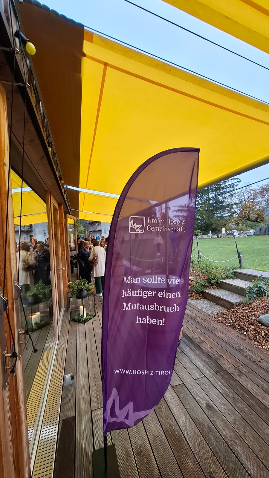 Eine lila Strandflagge der Tiroler Hospiz Gemeinschaft mit dem Spruch „Man sollte viel häufiger einen Mutausbruch haben!“ auf einer Holzterrasse unter einem gelben Sonnensegel.