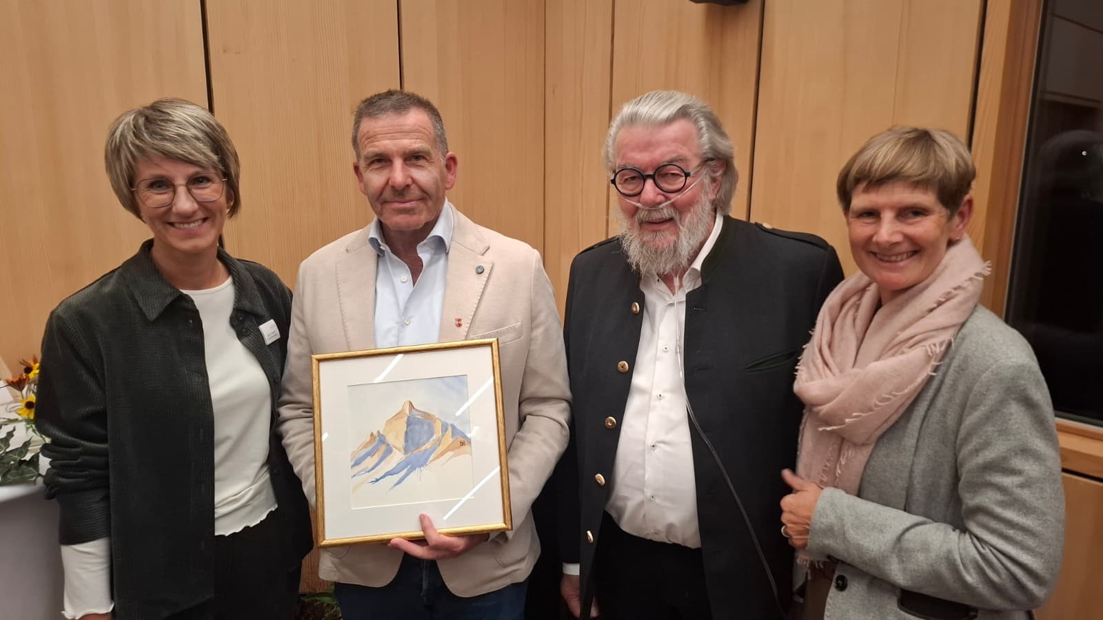 Vier Personen stehen nebeneinander vor einer Holzwand, während der zweite Mann von links ein gerahmtes Aquarellbild mit Bergmotiv präsentiert.