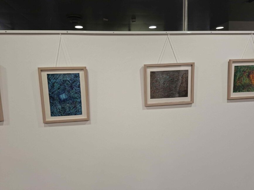 Aufnahme einer Kunstausstellung mit drei nebeneinander hängenden, gerahmten Kunstwerken an einer weißen Wand. Alle Bilder sind in hellen Holzrahmen mit Passepartout eingefasst und an Seilen von einer Galerieschiene abgehängt. Links ist ein hochformatiges Werk mit blau-grünen Mosaikstrukturen zu sehen, in der Mitte ein dunkles, feinstrukturiertes Bild im Querformat und rechts der Ansatz eines weiteren bunten, abstrakten Gemäldes.