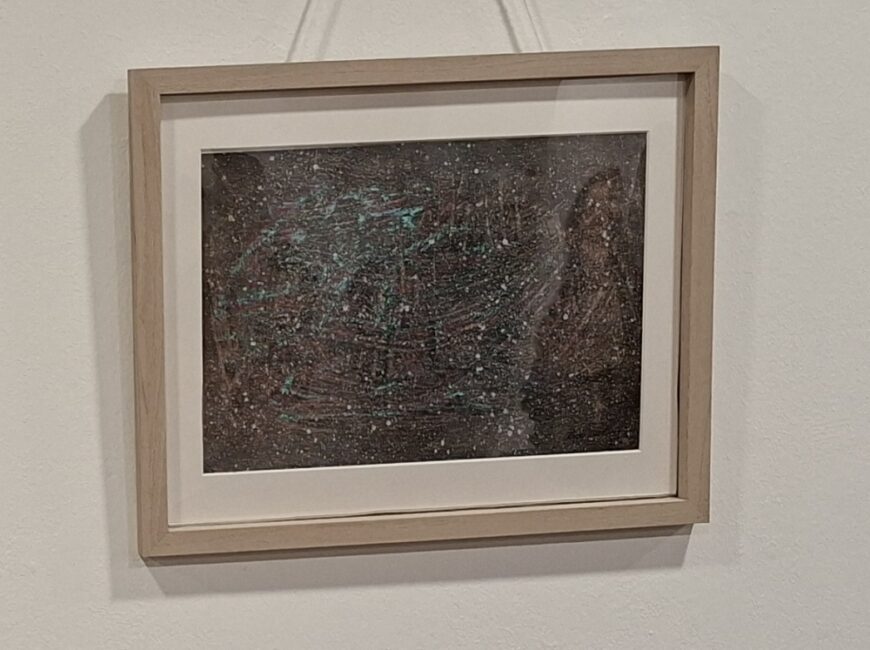 Ein gerahmtes, querformatiges abstraktes Gemälde an einer weißen Wand. Das Kunstwerk zeigt eine dichte, geschichtete Textur in Dunkelbraun, durchsetzt mit krakeligen hellblau-grünen Strichen und einer dichten Streuung von weißen Farbpunkten, die an eine Sternennacht oder fallenden Schnee erinnern. Es ist in einem schlichten, hellbraunen Holzrahmen mit weißem Passepartout gefasst und hängt an einer Schnur. Die Gesamtansicht ist leicht abgewinkelt, um die Tiefe des Rahmens zu betonen.