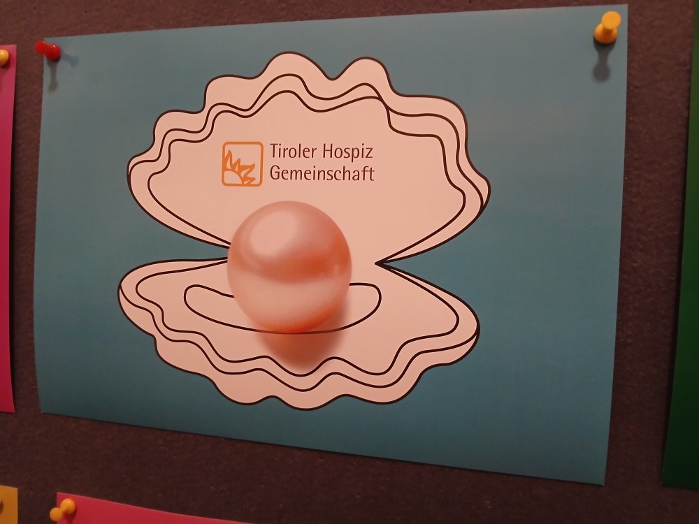 Eine blaue Karte an einer Pinnwand mit der Illustration einer geöffneten Muschel samt Perle und dem Logo der Tiroler Hospiz Gemeinschaft.