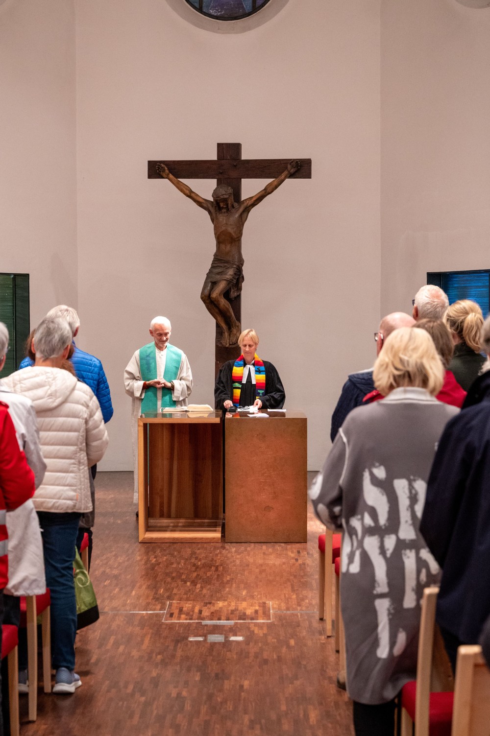 Blick durch den Mittelgang einer Kirche auf zwei Geistliche am Altar vor einem großen Kruzifix, während die Gemeinde stehend zuhört.