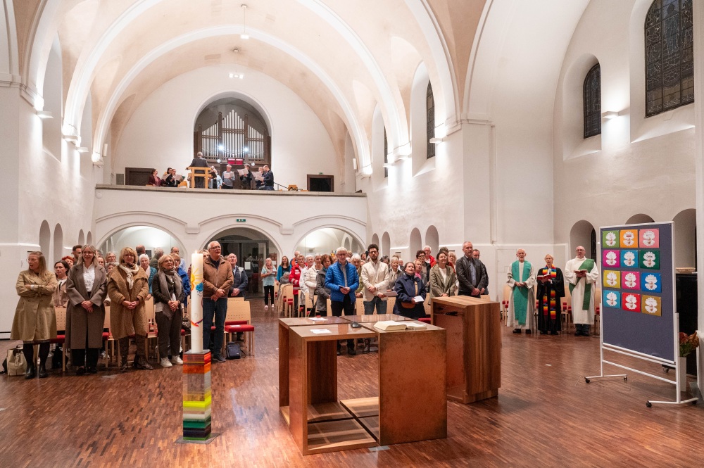 Panoramaaufnahme des Kirchenraums während eines Gottesdienstes mit stehender Gemeinde, dem Geistlichen-Team am Rand und dem Chor auf der Empore.