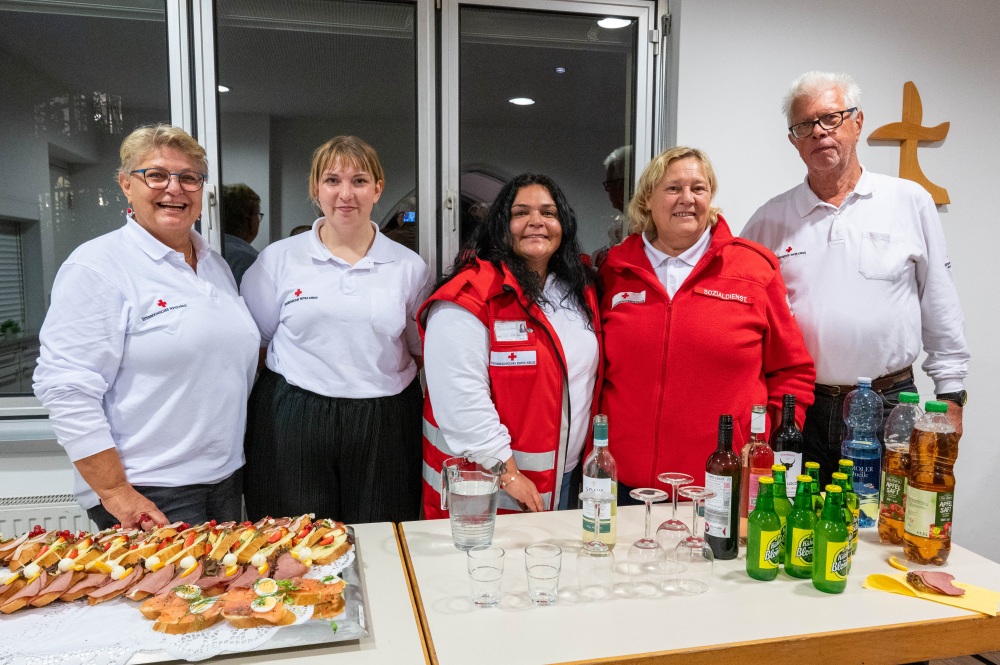 Fünf Helferinnen und Helfer des Roten Kreuzes stehen lächelnd hinter einem Buffet mit belegten Brötchen und Getränken.