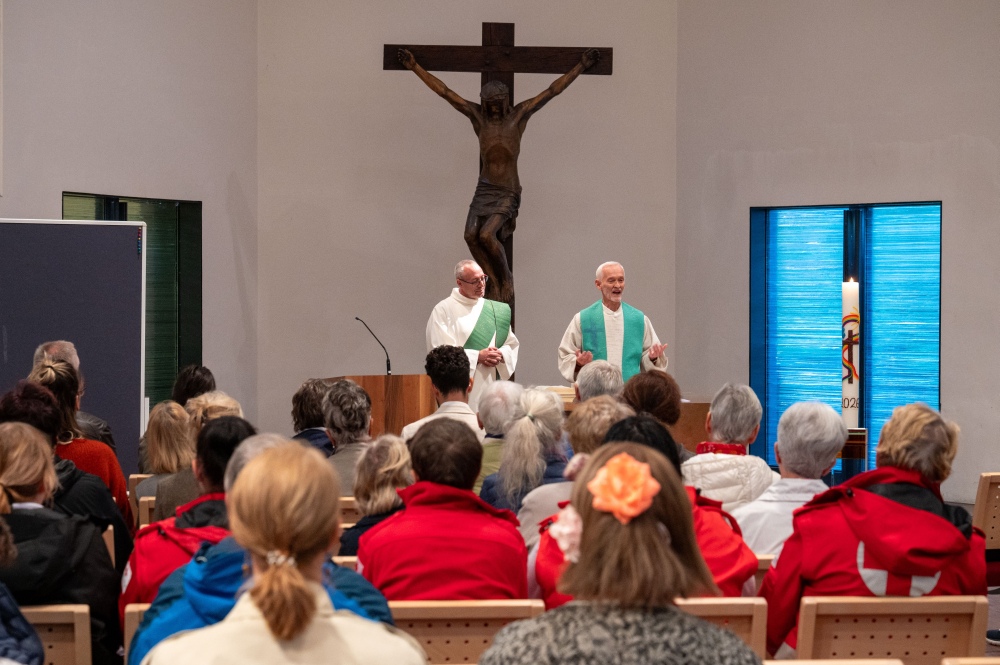 Ein Foto aus der Perspektive der Gemeinde während eines Gottesdienstes. Zwei Geistliche in weißen Gewändern mit grünen Stolen stehen vor dem Altar und sprechen zur Gemeinde. Im Hintergrund ist ein großes hölzernes Kruzifix an der Wand zu sehen.