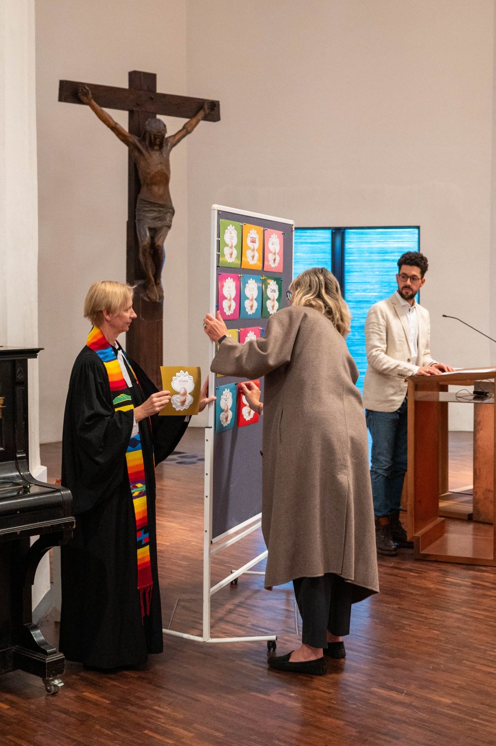 Eine Pfarrerin mit regenbogenfarbener Stola und eine Frau heften farbige Karten an eine Stellwand in einer Kirche.