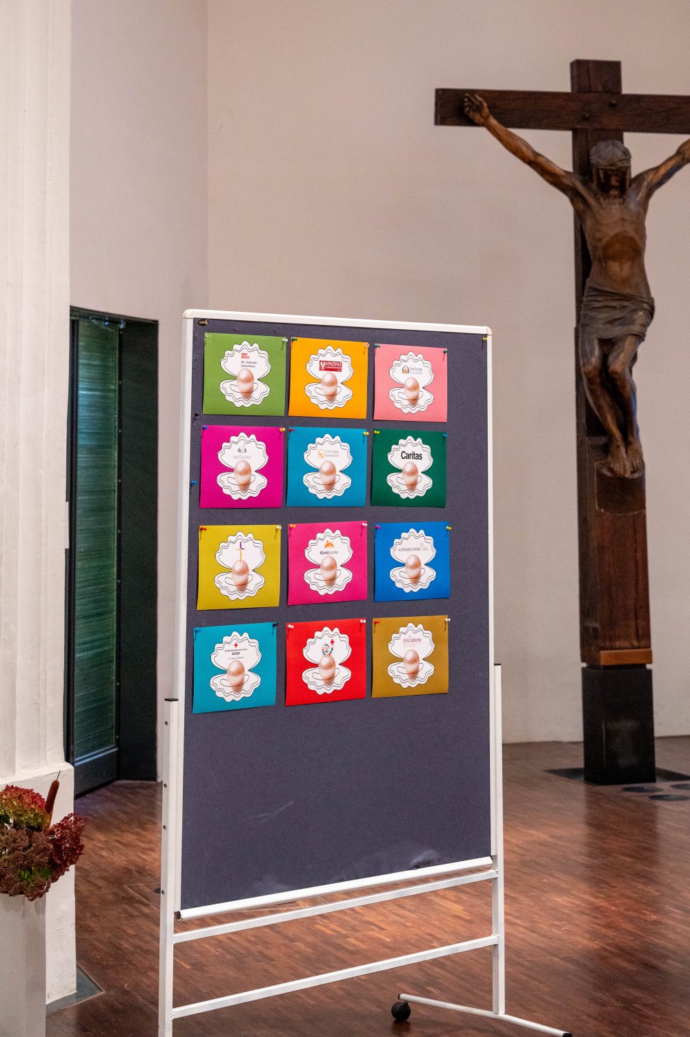 Eine mobile Stellwand in einer Kirche, an der zwölf bunt geordnete Karten mit Muschel-Motiven angepinnt sind, im Hintergrund steht ein großes Kruzifix.