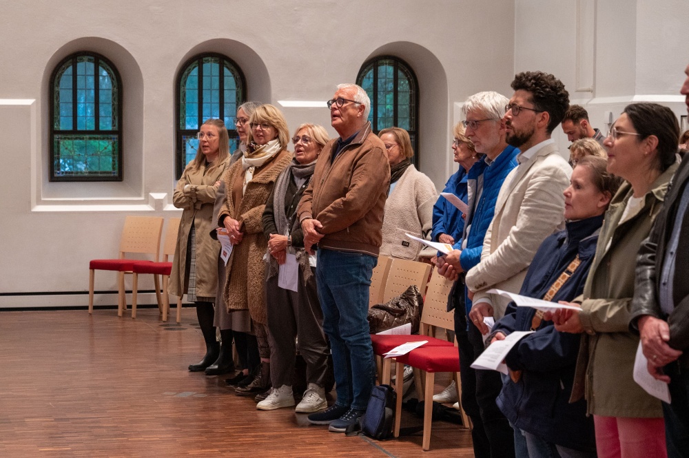 Stehende Gottesdienstbesucherinnen und -besucher in einer Kirche, die aufmerksam nach vorne blicken und teilweise Liederzettel halten.