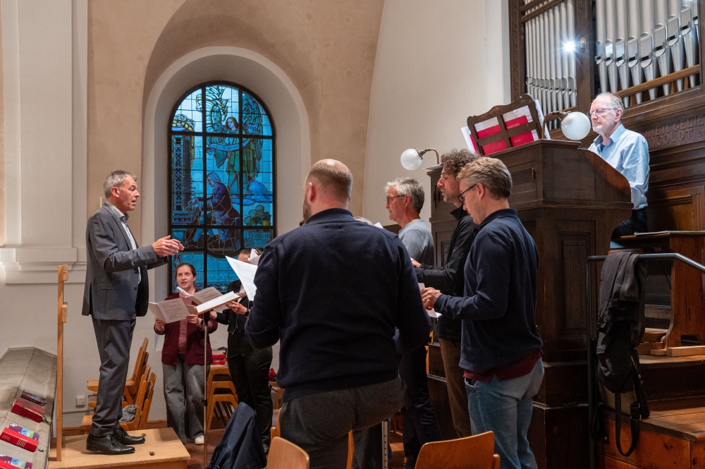 Ein Chor mit Chorleiter und Organist gestaltet die musikalische Begleitung eines Gottesdienstes auf der Empore vor einem bunten Bleiglasfenster.